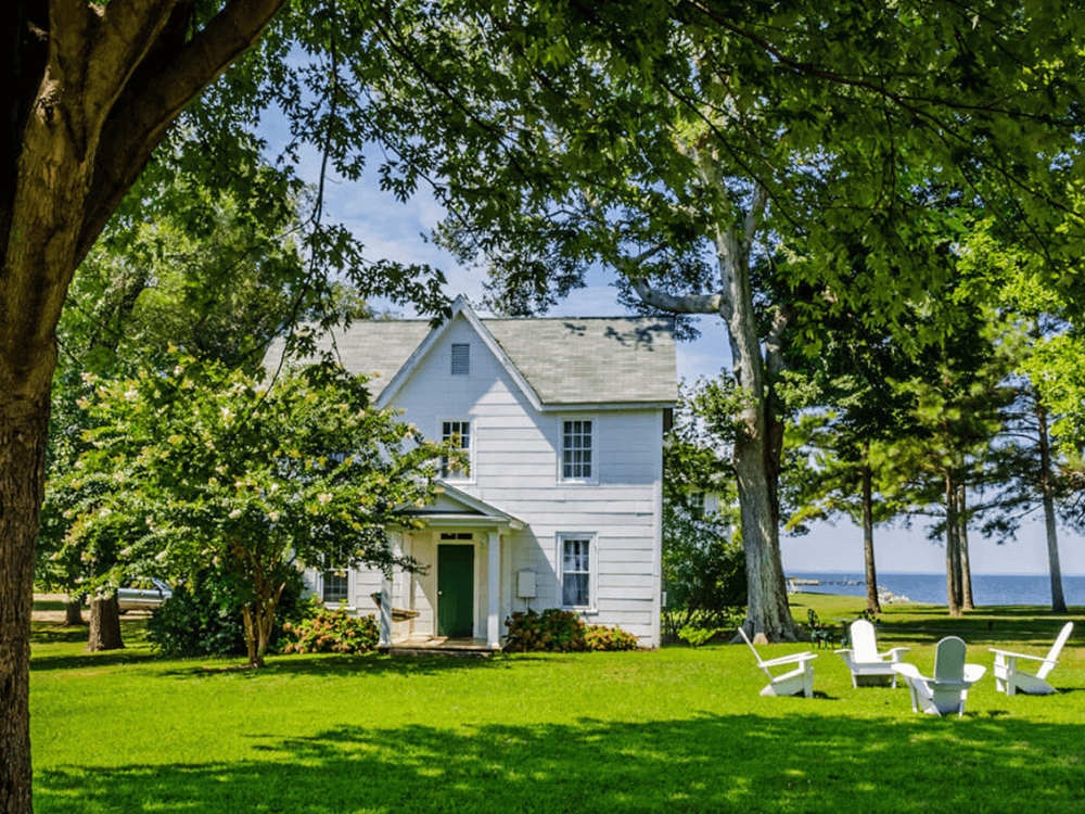A white house surrounded by green grass and trees, with adirondack chairs in the foreground and a view of the water in the background.